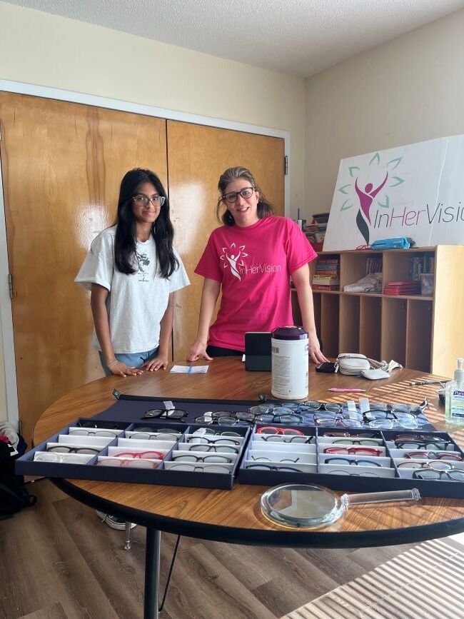 Two women standing behind a table lined up with various types of glasses frames.