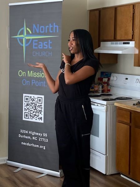 A lady talks to a group of people off camera while standing next to a banner that says North East Church. On Mission. On Point.