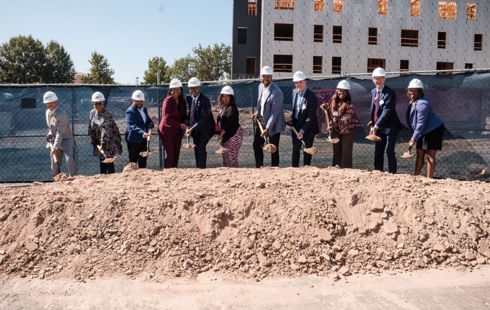 Eleven individuals wearing hard hats and each shoveling a pile of dirt for the groundbreaking of Dillard Apartments.