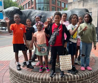 Eleven children smiling and waving in front of a bull statue.