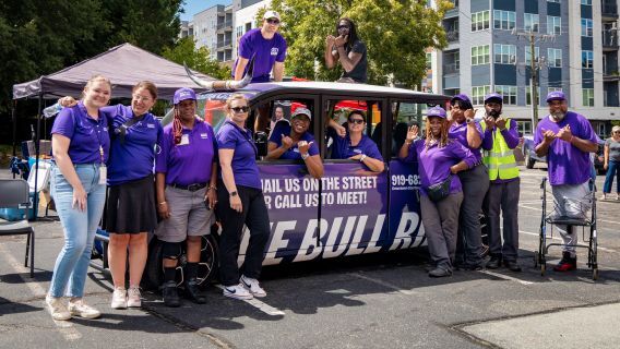 A group of individuals wearing purple while siting and standing around a purple car.