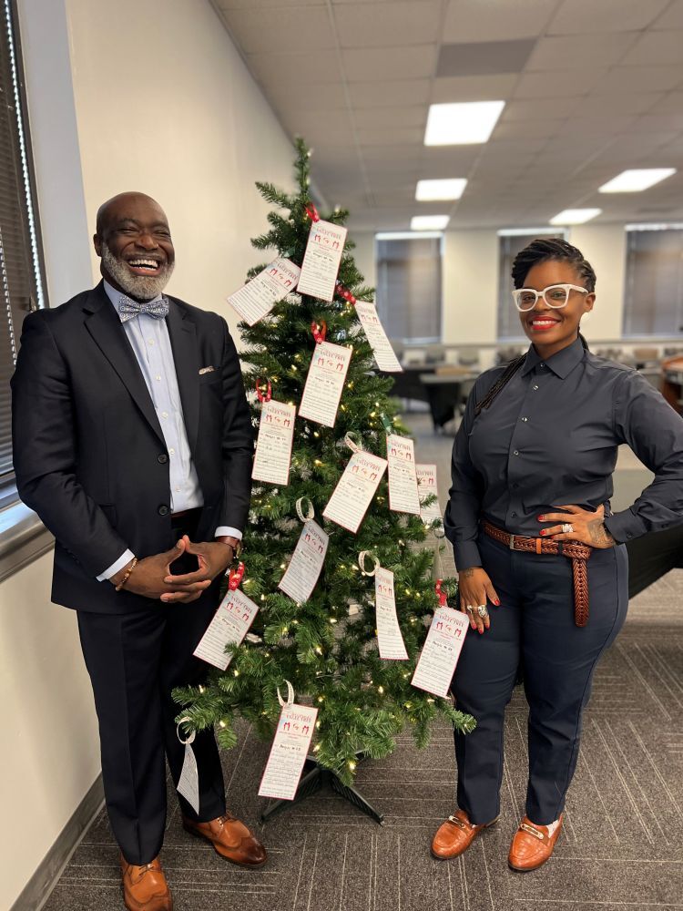 A man and a woman smiling while standing next to a Christmas Tree that has Angel Tree gift tags on it.