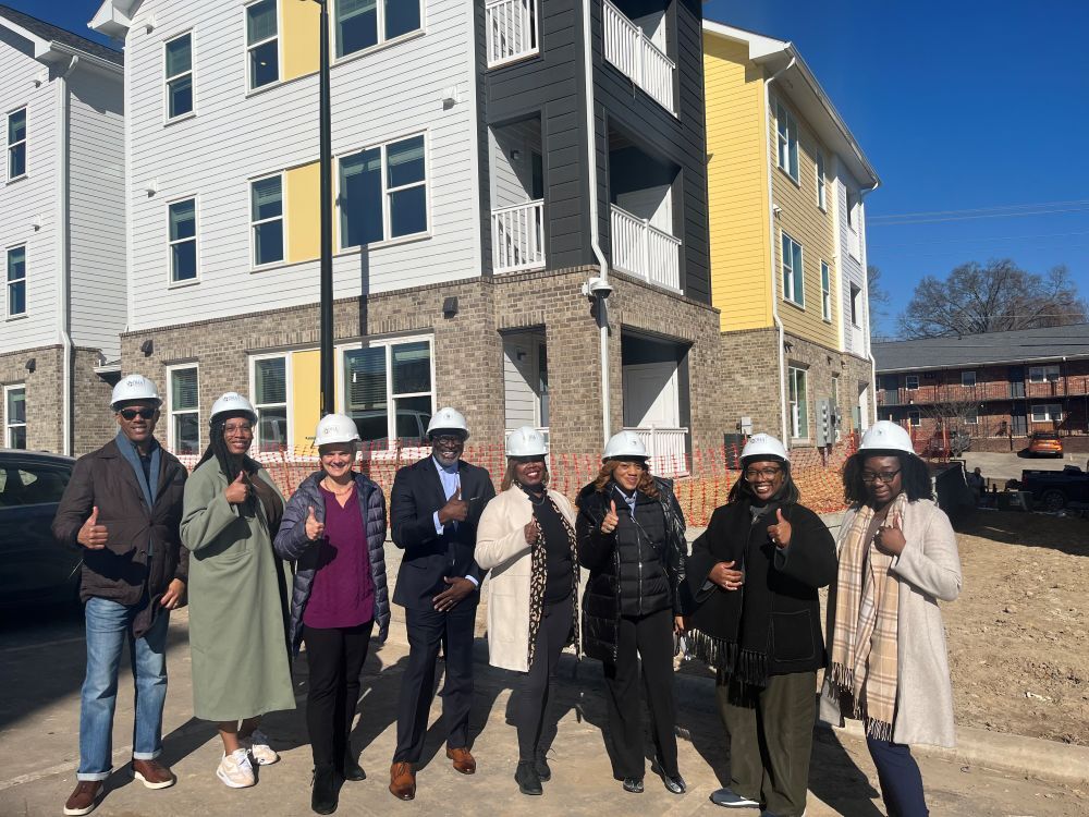 Eight individuals wearing hard hats, giving a thumbs up for the camera while they pose in front of a multi-story building.