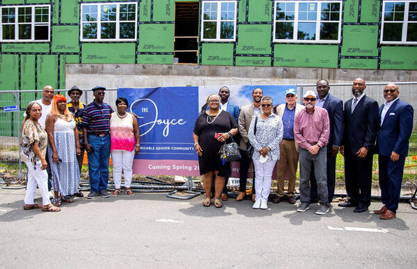 Several men and women are gathered around The Joyce construction site sign and smile.