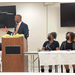 An individual standing at a podium next to two individuals sitting at a table.