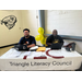 Two men sitting at a table with a table cover that reads: Triangle Literacy Council.