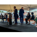 Two men stand and talk to a group of sitting individuals under a tent.